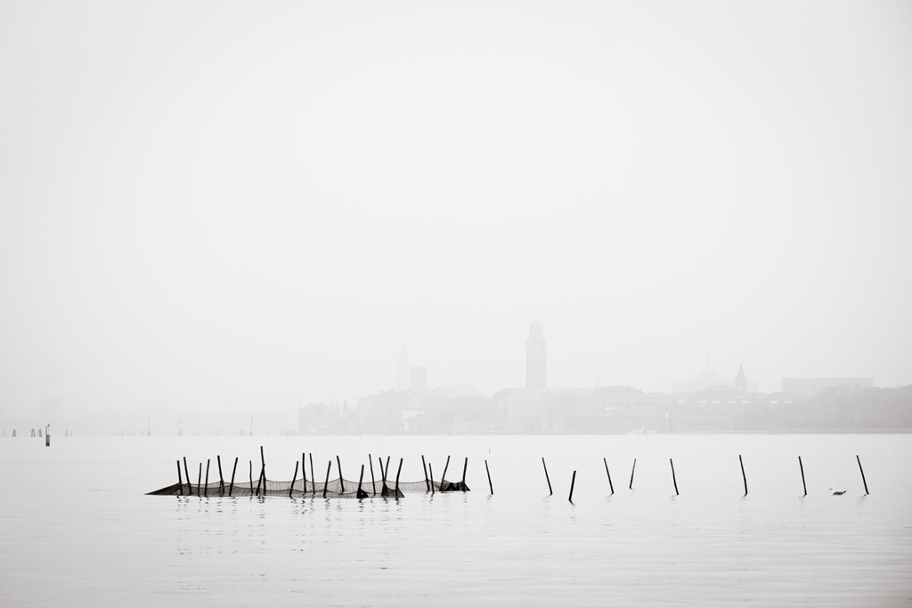 Venezia quasi dissolta nella nebbia con pali e reti in primo piano sulla laguna